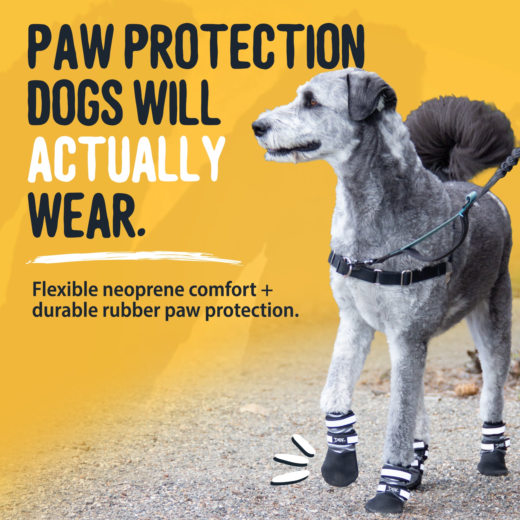 A gray dog wearing black paw boots and a black harness stands on a gravel surface against a bright yellow promotional background with large text reading 'PAW PROTECTION DOGS WILL ACTUALLY WEAR.' and smaller text about neoprene comfort and durable rubber paw protection.